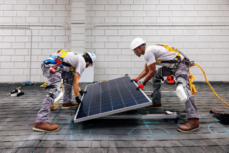 Two workers install a solar panel on a residential rooftop, wearing hard hats and safety harnesses