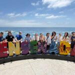Group of eleven people posing with colorful "Ensenada" letters at a coastal overlook with ocean and rocks in background