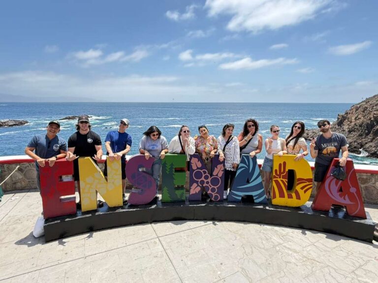 Group of eleven people posing with colorful "Ensenada" letters at a coastal overlook with ocean and rocks in background