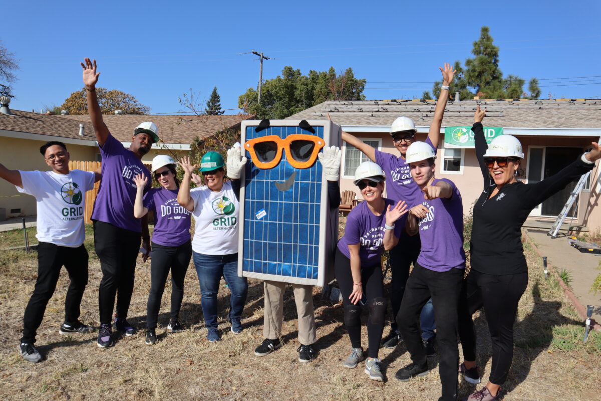 Team in hard hats and purple shirts celebrating with large solar panel and oversized glasses prop