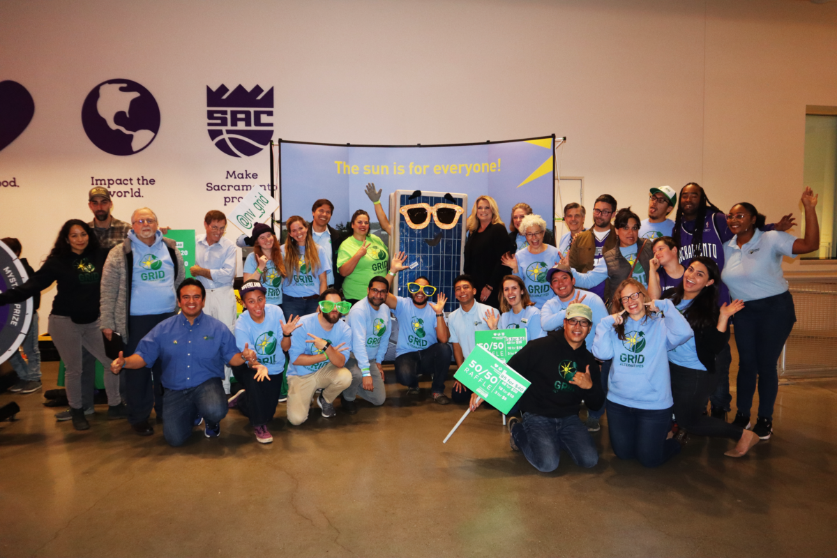 Large group of people wearing green and blue GRID shirts posing together indoors with solar panel banner, "The sun is for everyone!
