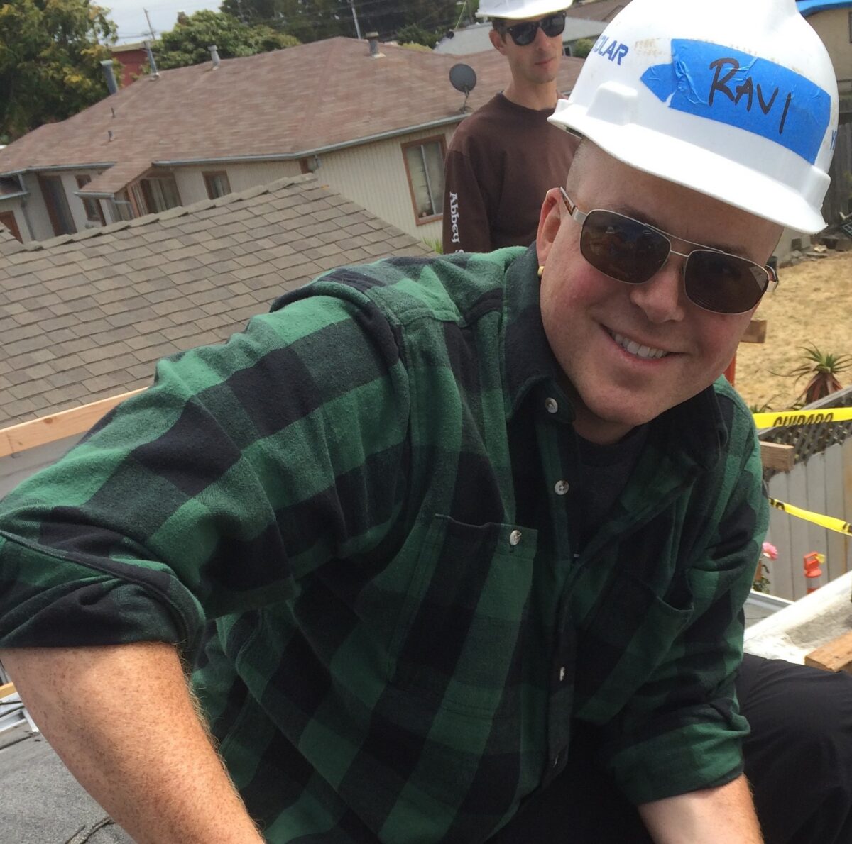 Man in green plaid shirt and white "Rani" hard hat on residential rooftop during construction work