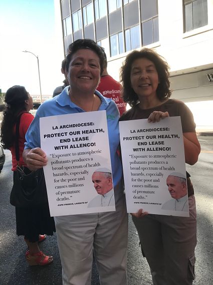 Two individuals supporting action by the Archdiocese hold up signs that say, "LA Archdiocese: Protect our health, end the lease with AllenCo"