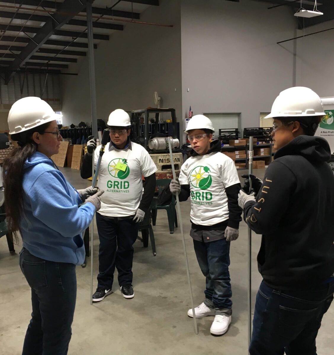 Five people wearing hard hats and work gloves stand in a warehouse, three wearing GRID Alternatives branded shirts during what appears to be a training or work session