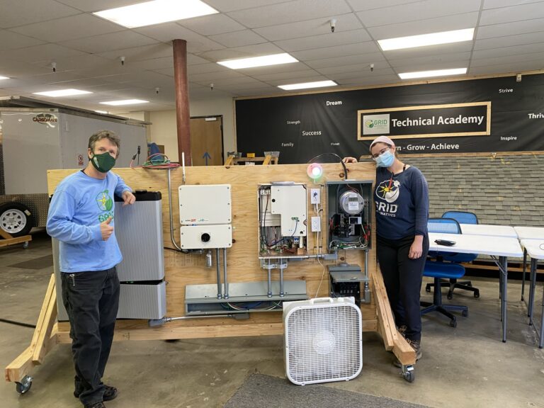 Two people displaying a wooden-framed HVAC system demonstration unit at GRID Technical Academy