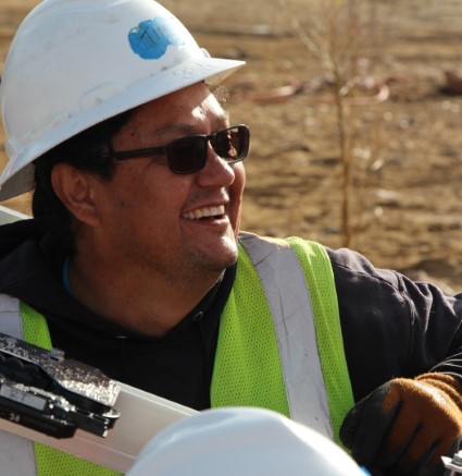 Construction worker wearing white hard hat, sunglasses, and green safety vest smiling outdoors
