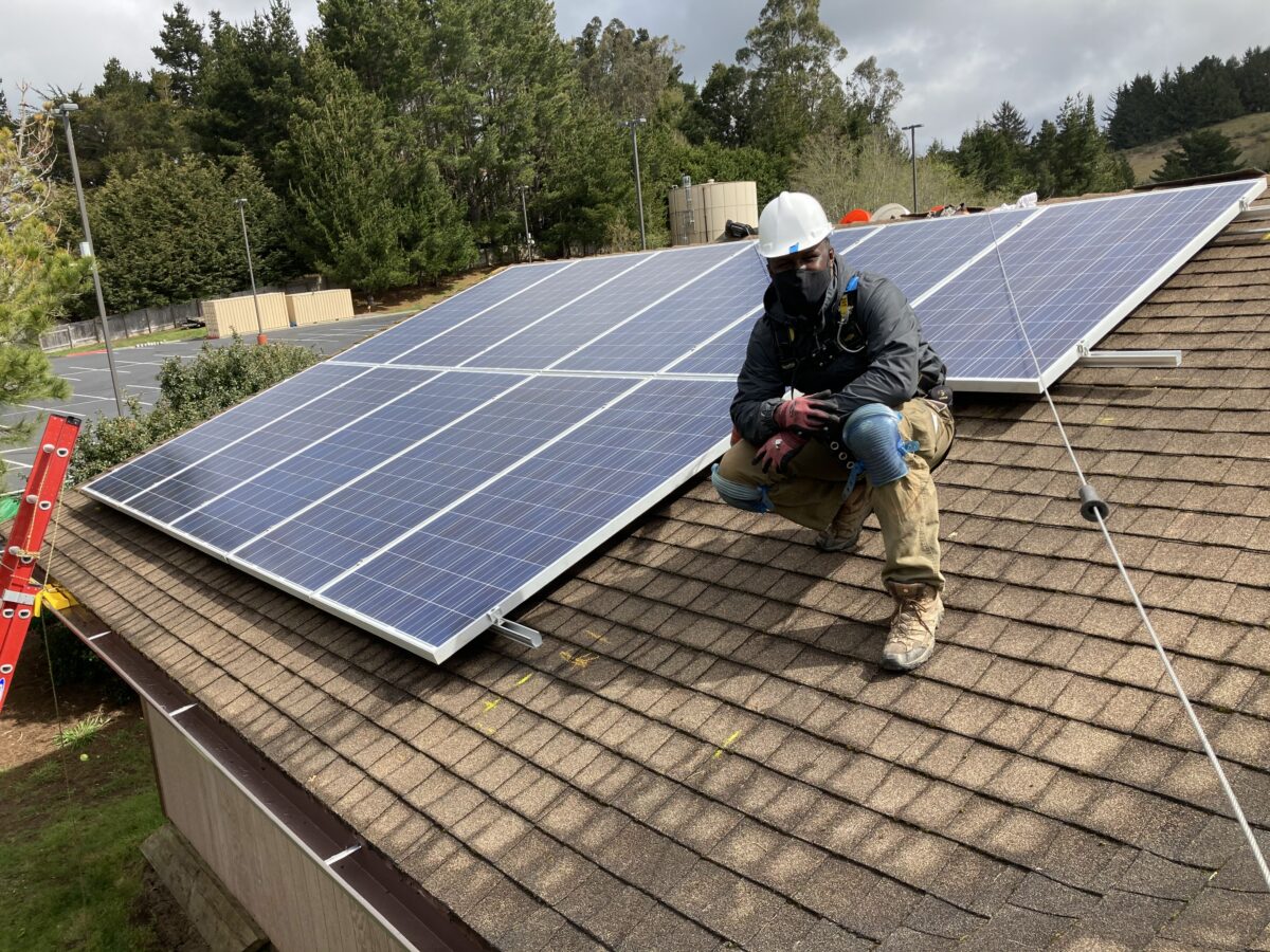 Worker in safety gear installing solar panels on residential roof with forest background