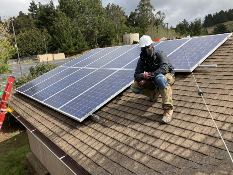 Worker in safety gear installing solar panels on residential roof with forest background