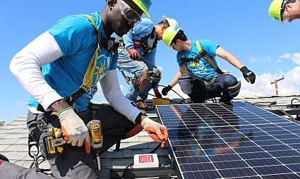 Three workers in safety gear installing solar panels on a rooftop under clear skies