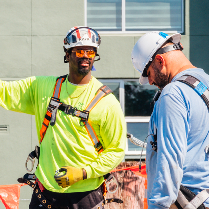 Two construction workers in safety gear discuss a project at a job site