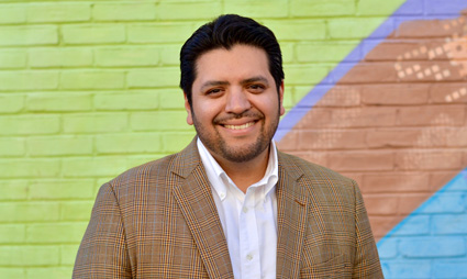 Professional headshot of man in tan blazer and white shirt against colorful brick wall