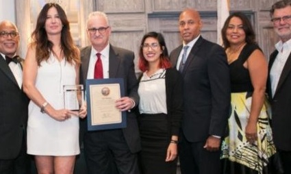 Seven people in formal attire posing together, one holding a framed award certificate