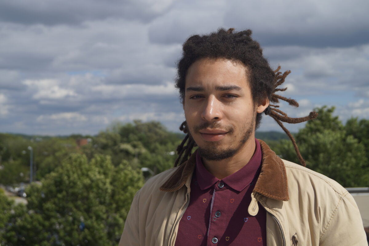 Man with dreadlocks wearing burgundy shirt and beige jacket standing outdoors with green landscape and cloudy sky behind him