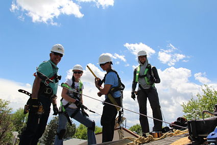 Four people in safety gear and hard hats stand on a wooden platform during an outdoor team-building activity
