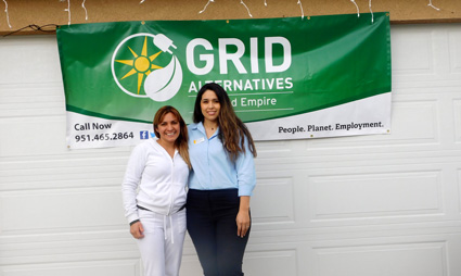 Two women standing under a green GRID Alternatives banner with contact information and "People. Planet. Employment." tagline