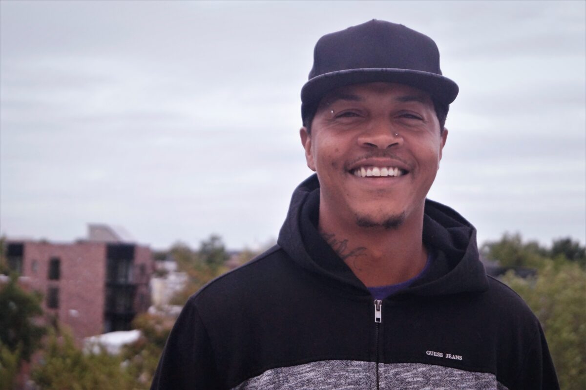 Smiling man wearing black Guess Jeans hoodie and cap, with urban buildings in background