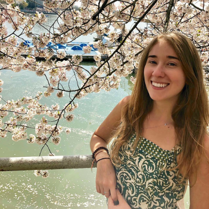 Smiling woman with long brown hair wearing a green patterned top, standing under cherry blossom branches with water visible behind her