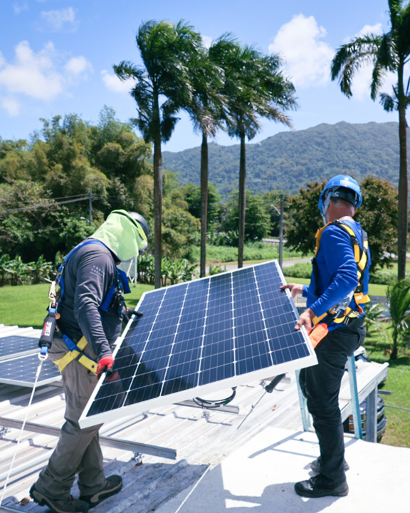 Two workers in safety gear installing a solar panel on a rooftop with mountains and palm trees in the background