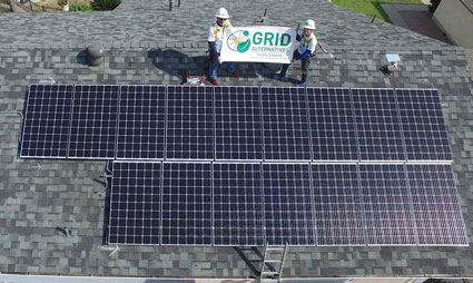Two workers hold a GRID banner while standing on a residential roof with two solar panel arrays installed
