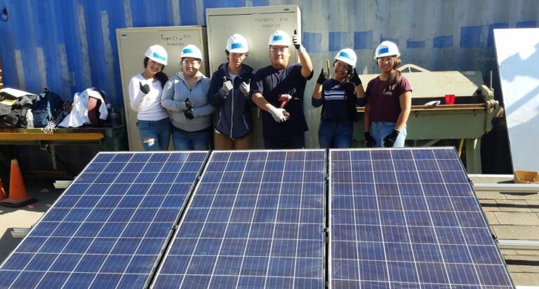 Six workers in hard hats pose behind installed solar panels on a roof