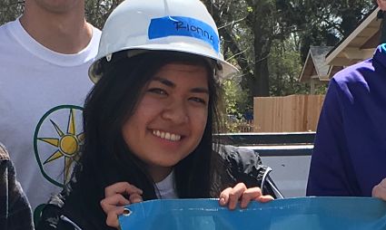 Woman in hard hat holding blue sign at outdoor event, smiling at camera