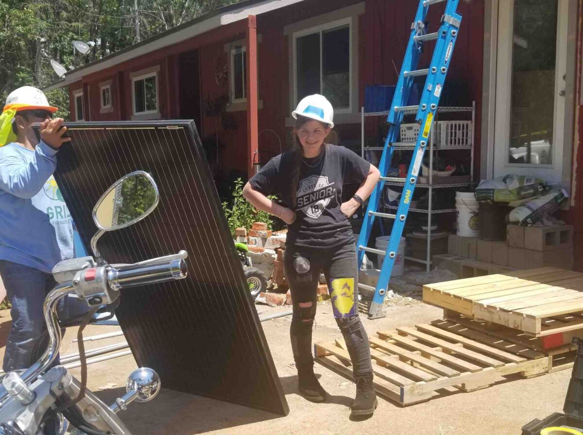 Young woman in hard hat standing by solar panel installation with workers and blue ladder at red house
