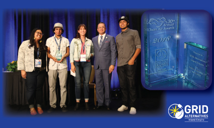 Five people and one man in business attire stand together at GRID Alternatives event, with blue digital display visible