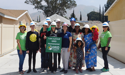 Group of volunteers in hard hats and colorful shirts posing with a solar panel installation sign on a residential roof