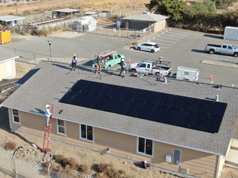 Solar panel installation on residential roof with work crew and vehicles in parking lot below