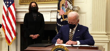 Man in suit signing document at Resolute desk with woman standing nearby; American flags visible