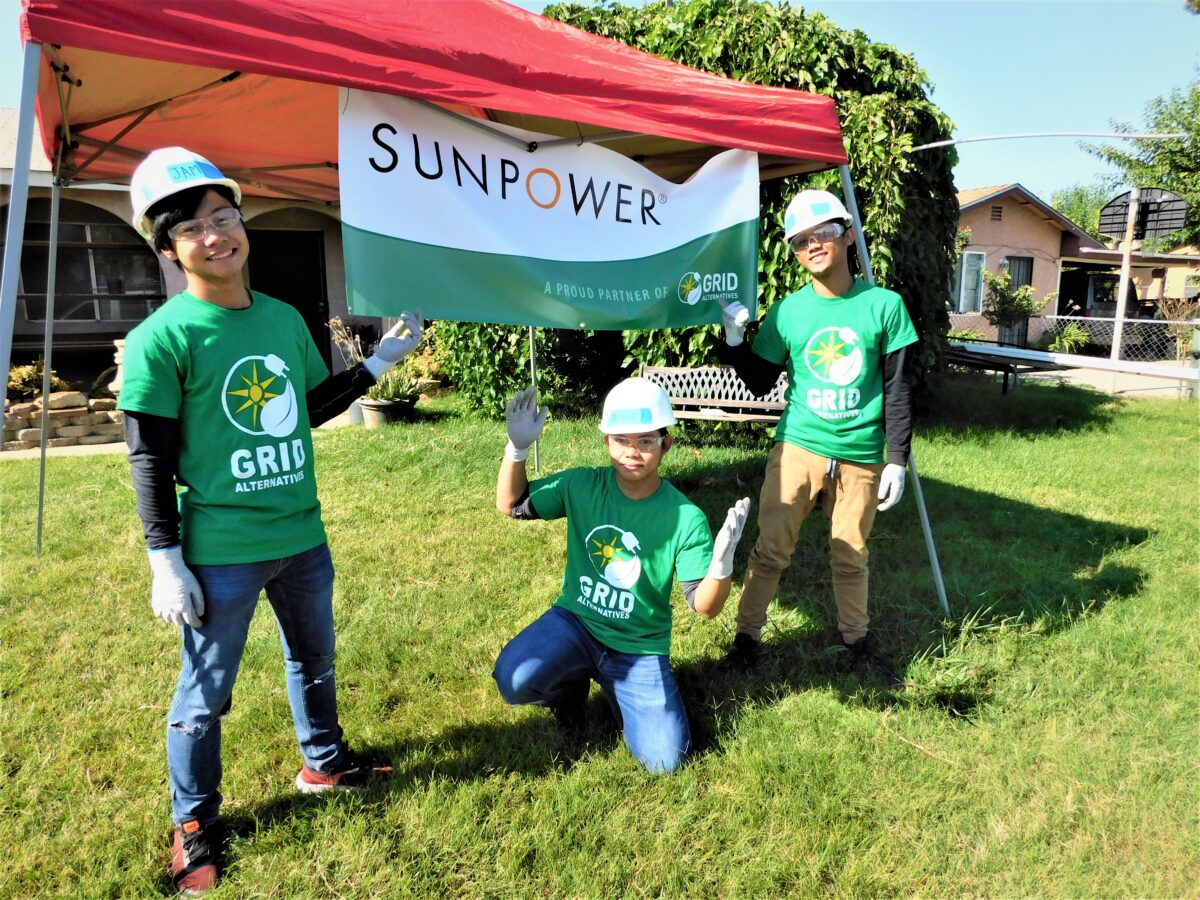 Three volunteers in green GRID shirts and hard hats hold a SunPower banner at an outdoor community event