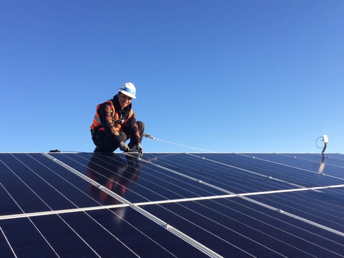 A trainee wearing an orange vest smiles while securing a solar panel on top of a roof. The sky is blue behind him.