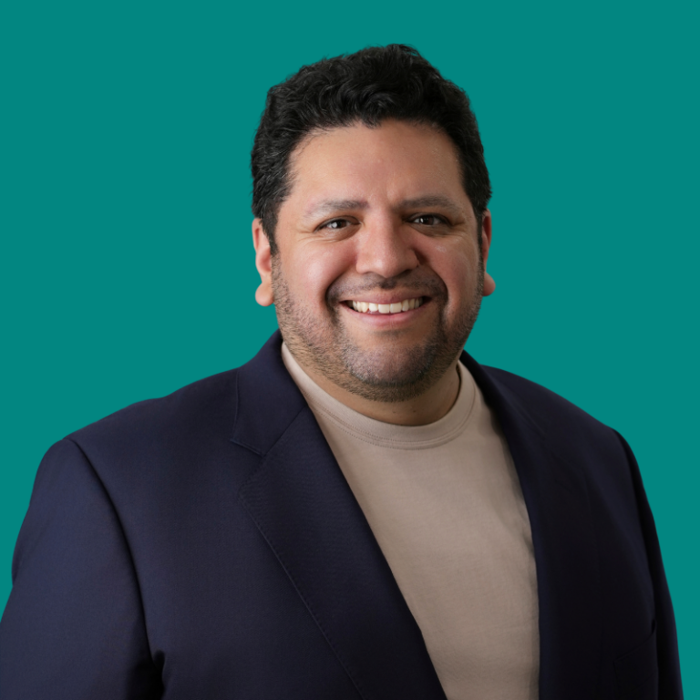 Jaime Alonso, man smiling at camera wearing navy blazer and beige shirt against teal background
