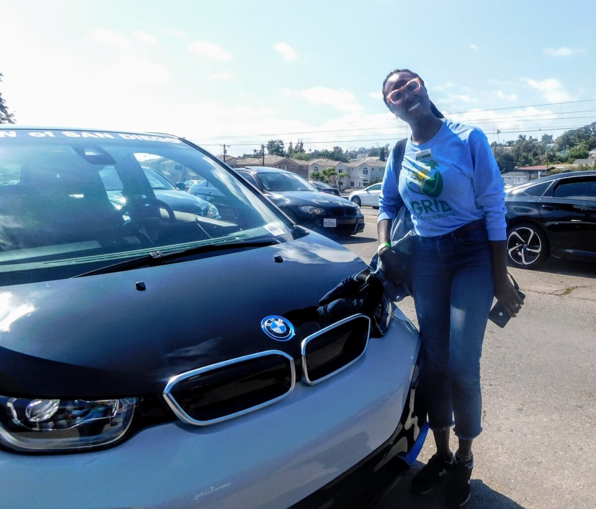 Person in blue "Grid" sweatshirt posing with white BMW i3 electric vehicle at outdoor event
