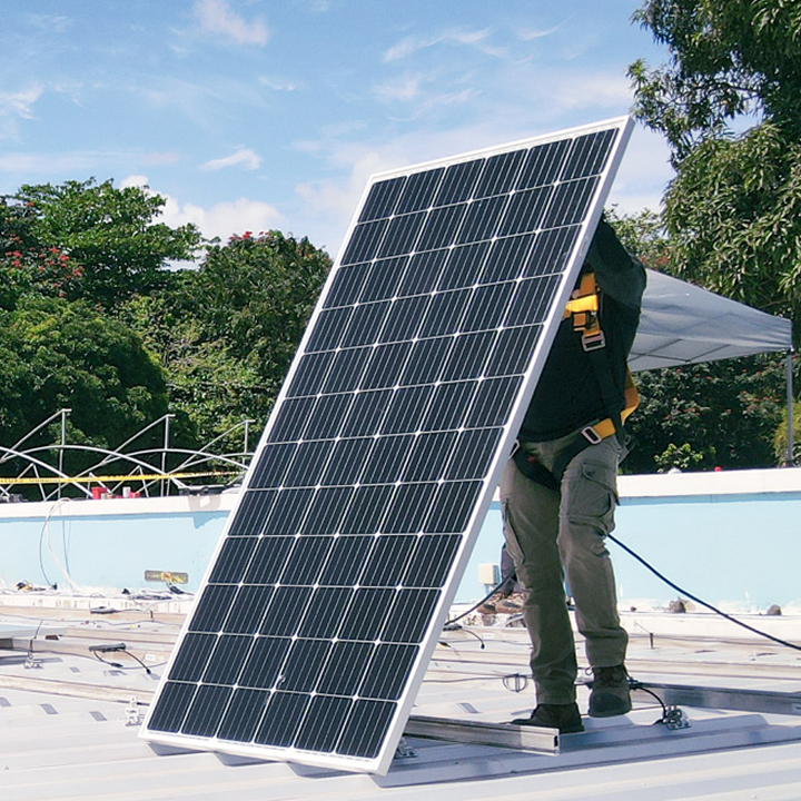 Worker installing a large solar panel on a flat roof with blue sky and green trees in background