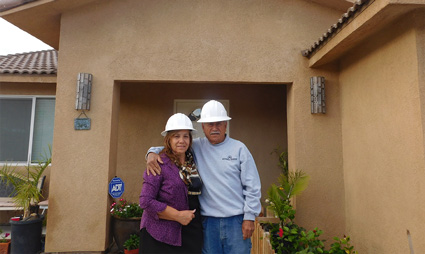 Two people wearing hard hats stand in a home entrance doorway, smiling at the camera