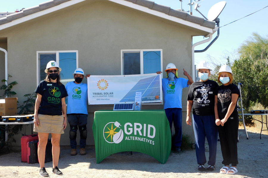Six people in hard hats and masks stand behind a green GRID Alternatives table with solar panel display in front of a residential house