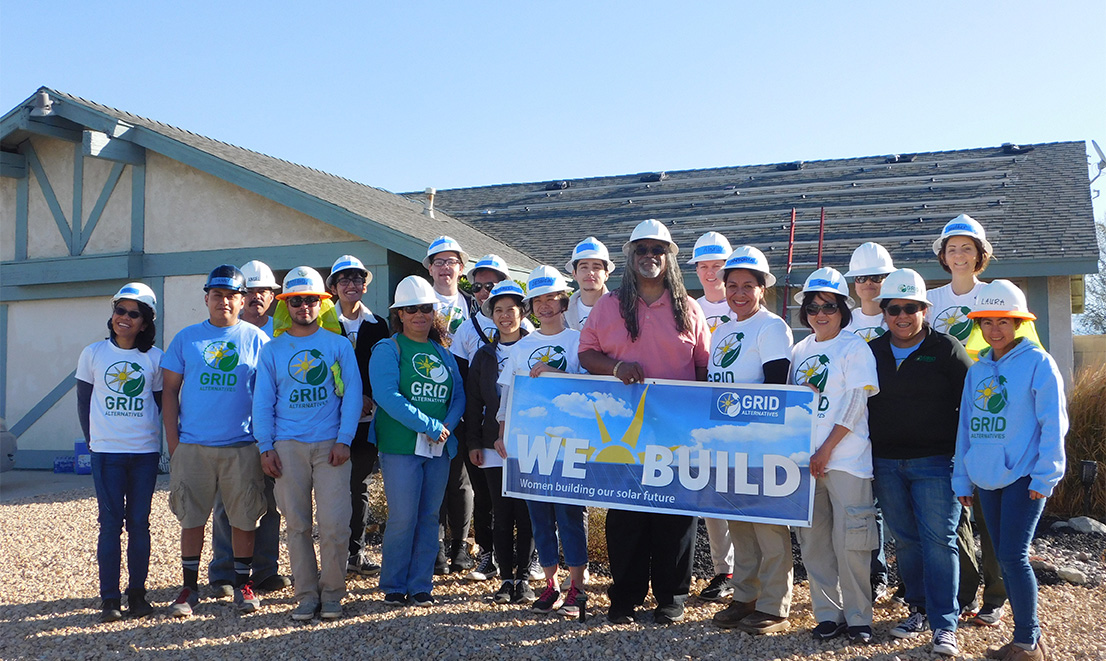 Group of solar installation workers wearing hard hats and GRID shirts holding "We Build" banner in front of house