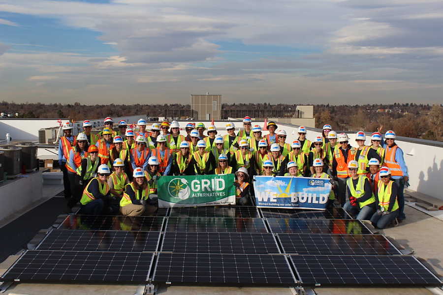Large group of construction workers in safety gear on rooftop with solar panels, holding "Grid Alternatives" and "We Build" banners