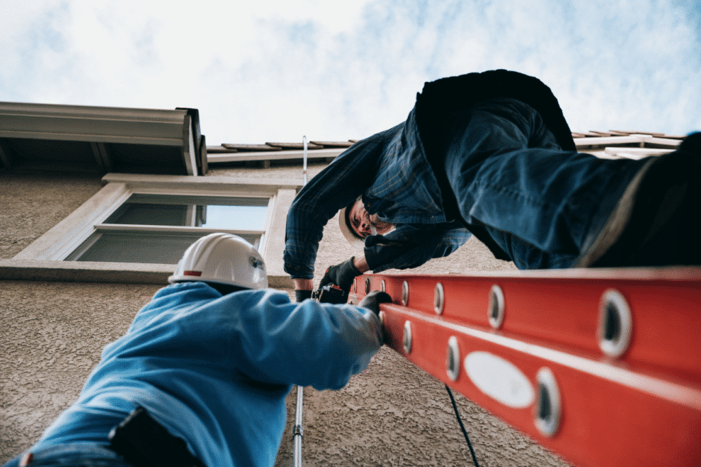 Seen from the ground, one installer steadies a red extension ladder while another leans over the roof edge.