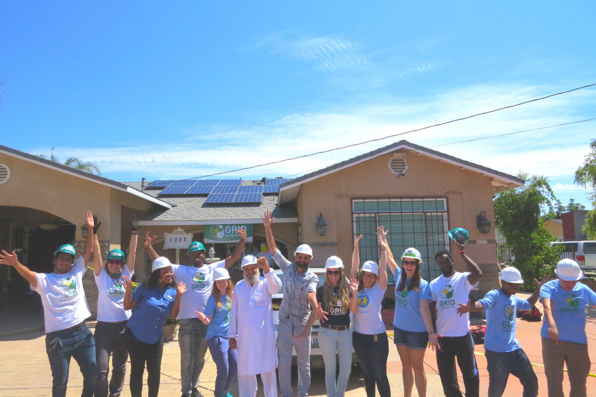 Group of people in hard hats and GRID t-shirts celebrating in front of a house with solar panels