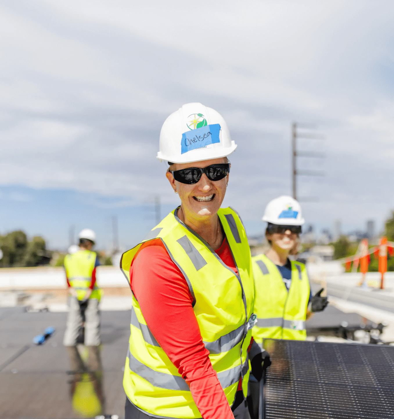 A GRID installer in a hard hat and safety vest smiles over her shoulder while working on a rooftop solar array.