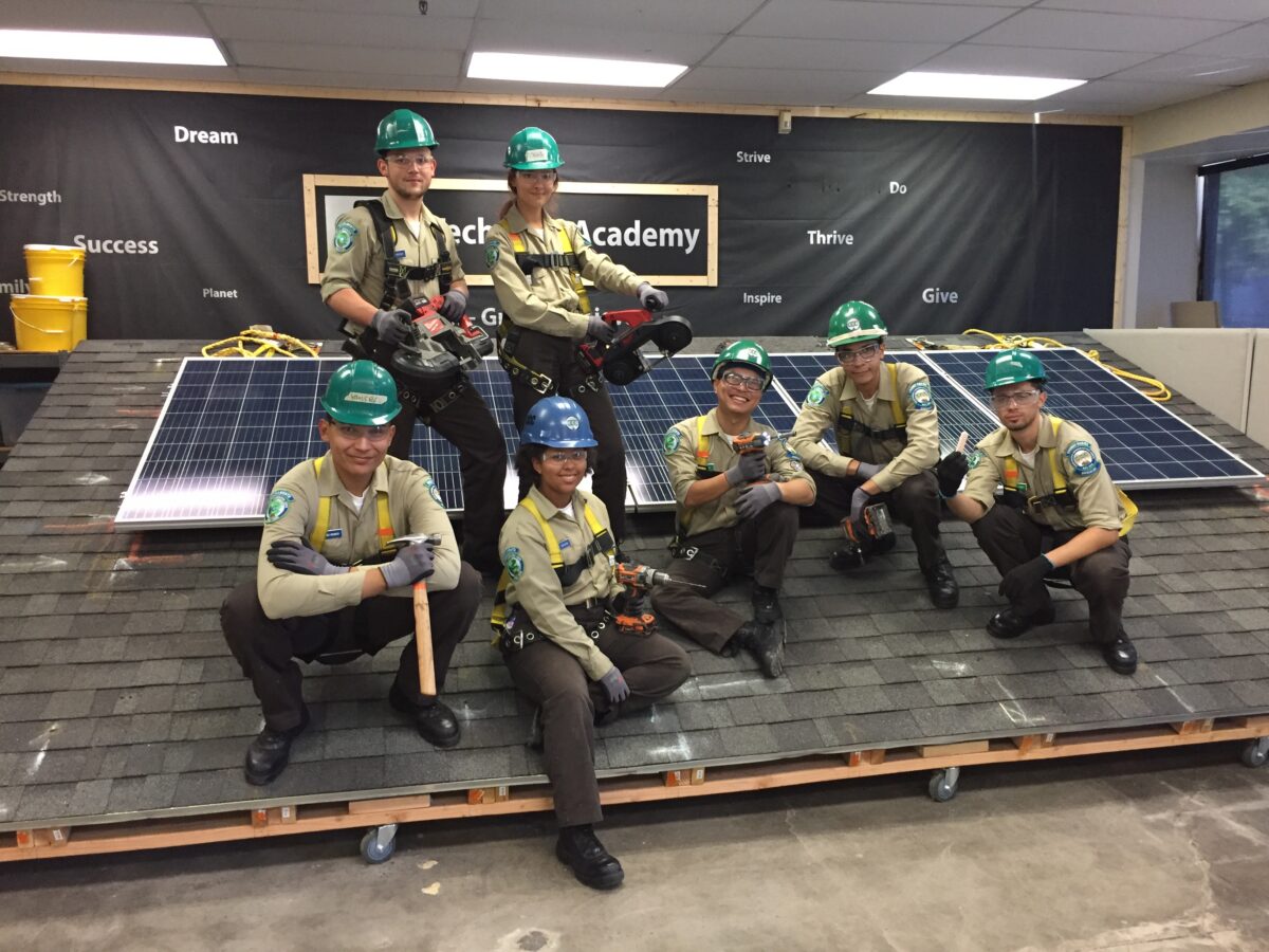 Seven scouts in hard hats and uniforms posing on solar panels at Ceck Academy with motivational words on the backdrop behind them