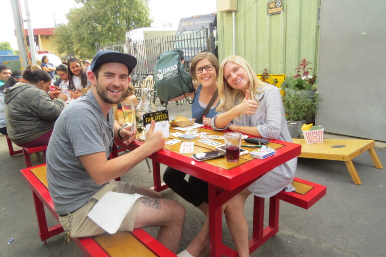 Three people smiling at a red picnic table with drinks and cards at an outdoor cider event