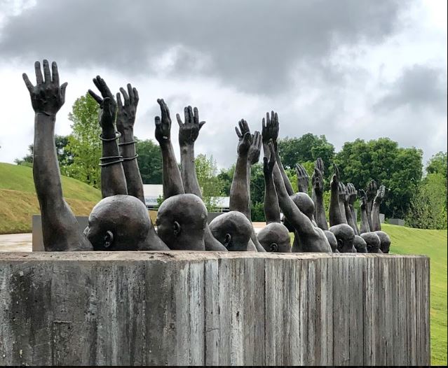 Bronze sculpture of emerging hands and heads rising from concrete base in grassy landscape