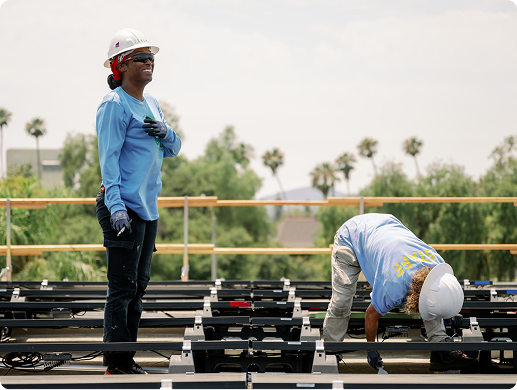 Two construction workers installing solar panels on a rooftop with palm trees in background