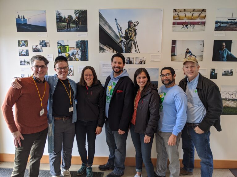 Seven staff members smiling at camera in front of wall displaying photographs and project documentation