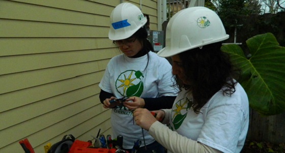 Two workers in hard hats and matching green shirts inspect exterior house siding with tools