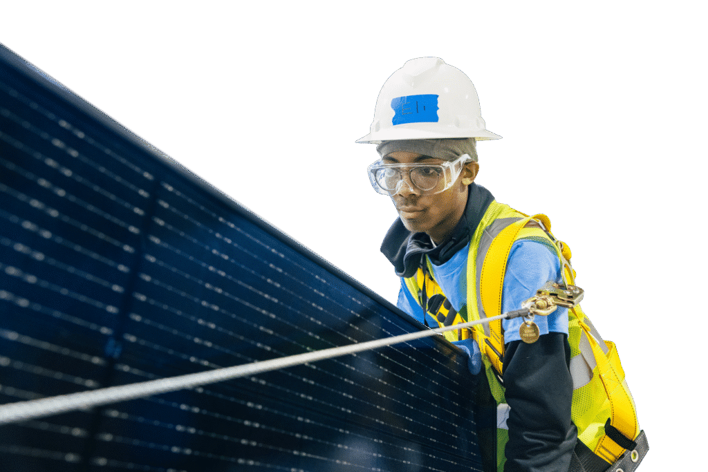 A trainee in a hard hat, safety goggles and a fall-arrest harness guides a solar panel into position.