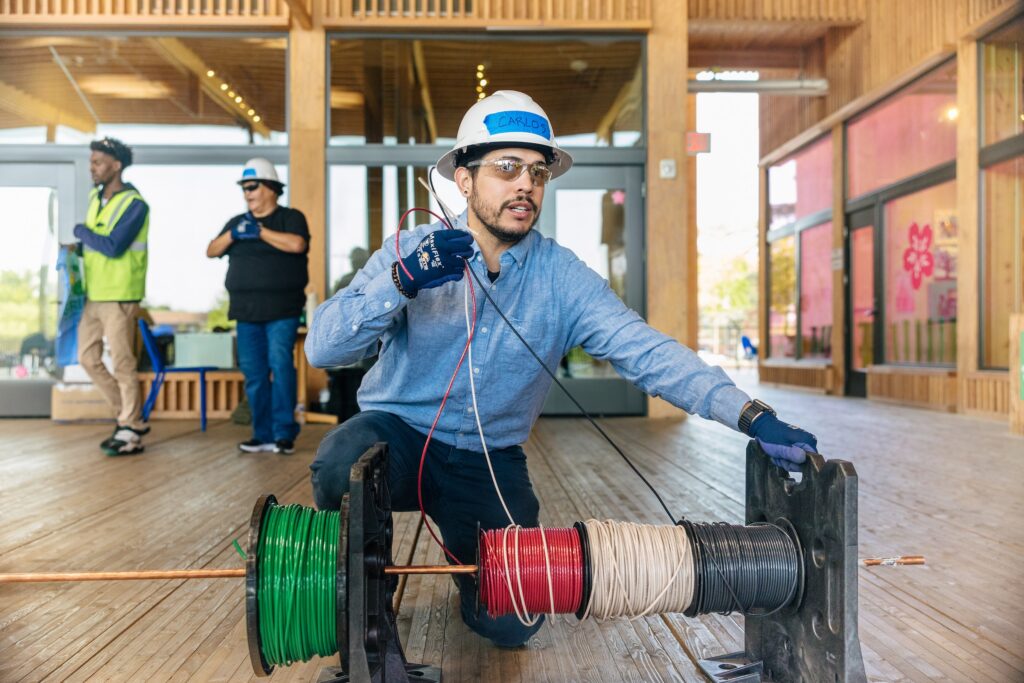 A trainee carries two large spools of electrical cable across a training bay.